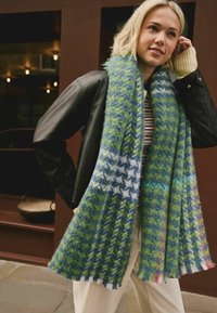 Ung kvinde iført et stort grønt og blåt houndstooth-scarf, sort læderjakke, stribet skjorte og cremede bukser i en urban setting.