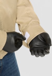 Svetlo rjav plašč z gladko teksturo, zadrgo in belim Velcro trakom na rokavu. Črne lateks rokavice so oblečene na obeh rokah.