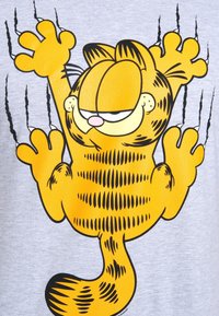 Camiseta gris con un gato de dibujos animados, de color naranja con rayas negras. El gato está representado trepando con las garras extendidas sobre un fondo texturizado.