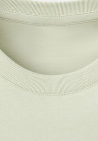Close-up van een lichtbeige ronde hals met zichtbare geribbelde stiksels op een effen katoenen t-shirt.