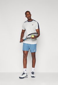 Hvitstripete t-skjorte med marineblå detaljer, blå shorts, holder et racket og en ball. Sportssko og sokker med logo synlige. Enkel bakgrunn.