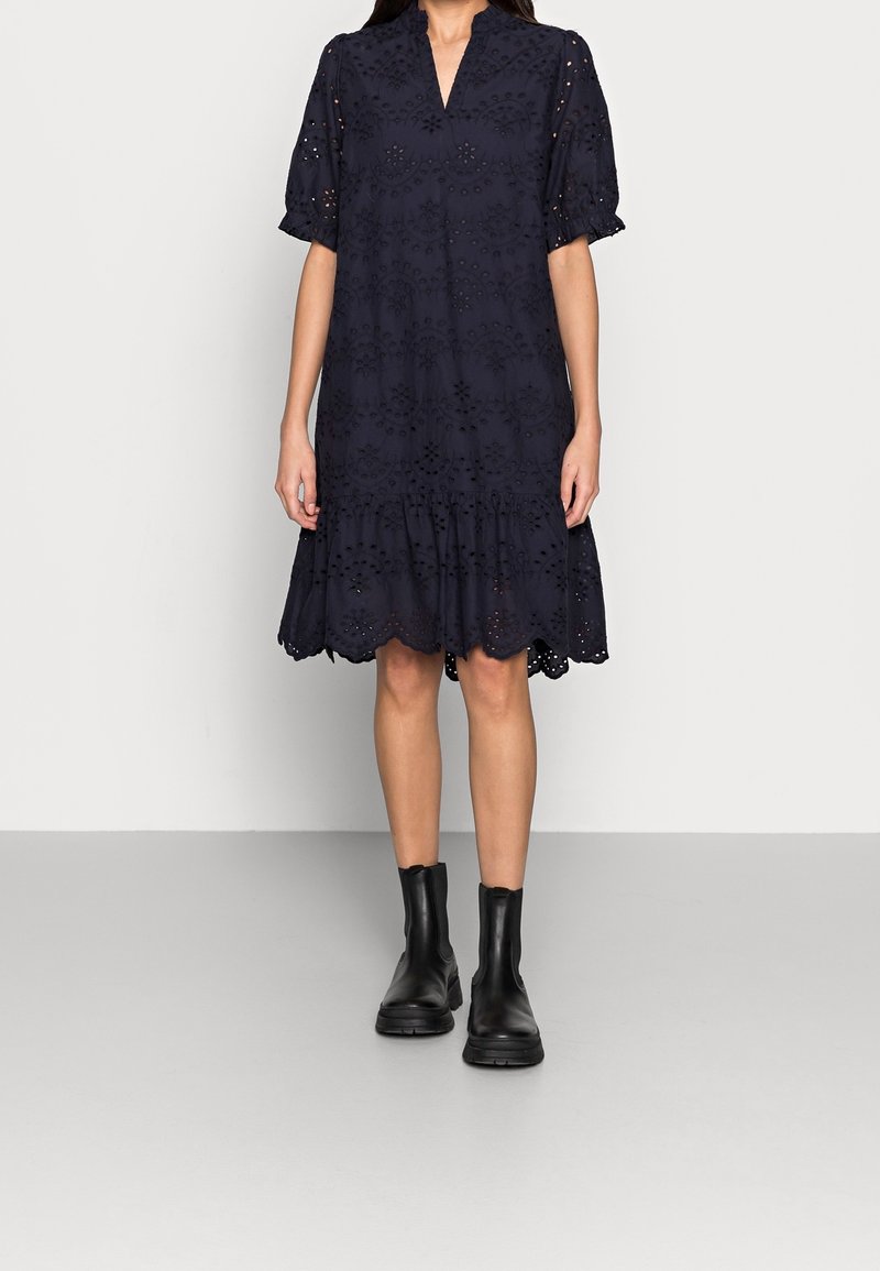 Robe avec des manches courtes bleu marine, ornée de broderies, décolleté en V et ourlet à volants. Portée avec des bottines noires sur un fond gris clair.