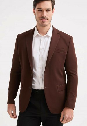 Jeune homme portant un blazer marron sur une chemise blanche et un pantalon noir, debout devant un fond clair uni.
