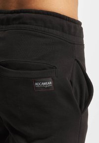 Melna Roca Wear sporta bikses ar elastīgu jostu, sānu kabatām un redzamām logotipa etiķetēm ar sarkaniem akcentiem. Mīksta auduma tekstūra.
