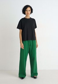 Zwarte katoenen T-shirt met korte mouwen, gecombineerd met losse, felgroene broeken. Het model staat in platte zwarte schoenen tegen een effen achtergrond.