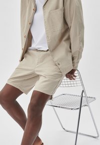 Beżowa, lekka bawełniana koszula z kieszonką, w połączeniu z pasującymi beżowymi szortami. Model siedzi na krześle z drutu. Styl minimalistyczny.