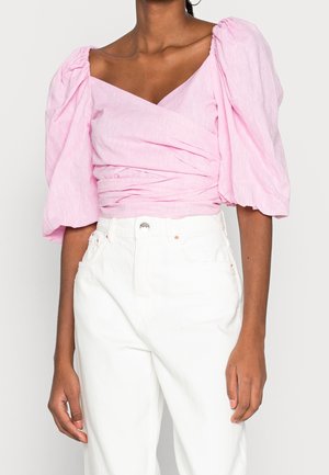 Frau trägt eine rosa Wickelbluse mit Puffärmeln und weiße, hoch taillierte Hose vor einem schlichten hellen Hintergrund.