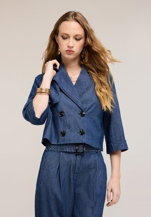 Kvinde med langt blondt hår iført en dobbeltknappet cropped denimjakke og højtaljede denim bukser med et flettet bælte.