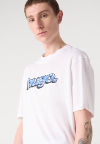 White cotton t-shirt featuring blue patterned graphic "hugo" text on the front. Short sleeves with a relaxed fit and crew neckline.