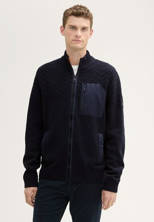 Navy zip-up sweater with textured zigzag design on the chest, smooth nylon pocket, and ribbed cuffs and hem.