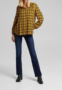 Camicia a maniche lunghe gialla e nera a quadri, vestibilità rilassata, con colletto button-down. Indossata con jeans a vita alta blu scuro e stivaletti neri.