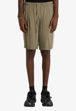 Persona che indossa pantaloncini beige con coulisse, calzini neri e sneakers nere chunky, in piedi con le mani rilassate lungo i fianchi.
