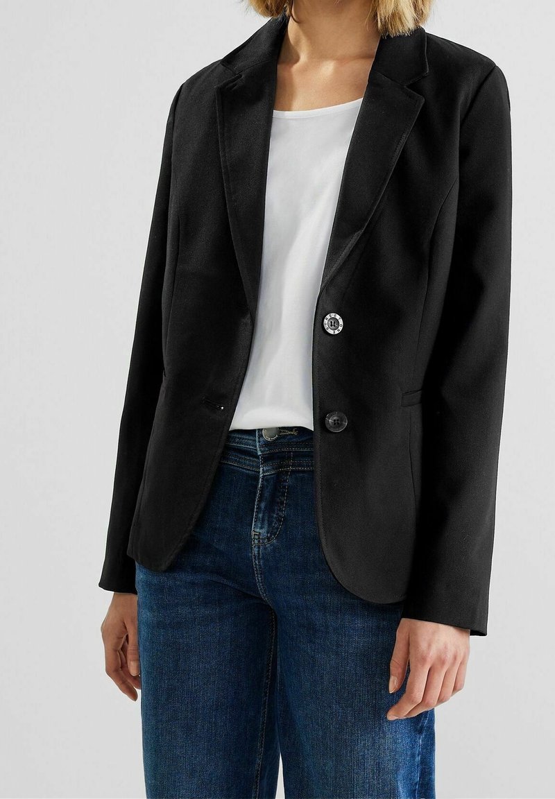 Femme portant un blazer noir ajusté sur une chemise blanche, associée à un jean en denim bleu, debout devant un fond clair uni.