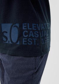 Donker marineblauwe katoenen T-shirt met een blauwe grafische ontwerp met "s.O." en de tekst "ELEVATED CASUAL EST. 1969" gedrukt op de zijkant.