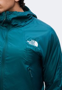 Άτομο με μια ελαφριά γαλάζια-πράσινη (teal) ζακέτα North Face με κουκούλα, φερμουάρ μπροστά και τσέπη, κοιτάζοντας αριστερά σε απλό φόντο.