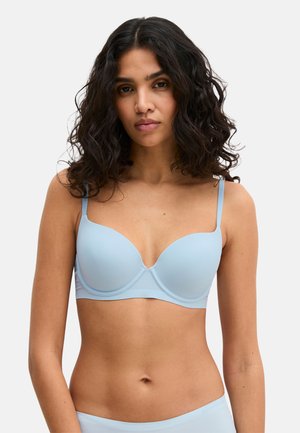 Vrouw met golvend donker haar, gekleed in een lichtblauwe gewatteerde beha en bijpassend ondergoed tegen een effen lichte achtergrond.