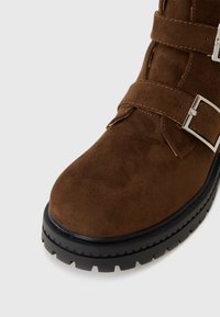 Bota de ante marrón hasta el tobillo con hebillas plateadas, punta redonda y suela de goma negra gruesa con borde ondulado.