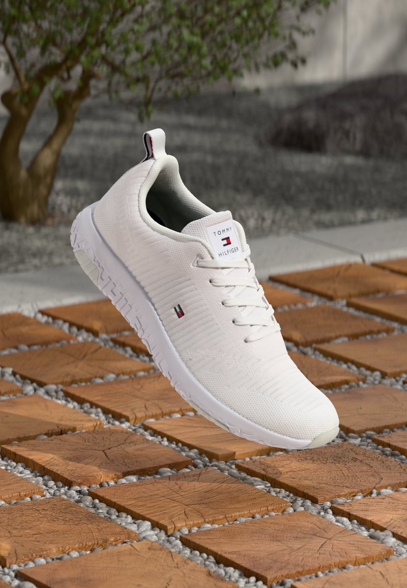 Chaussure de sport blanche avec tissu texturé, système de laçage, un écusson logo et une semelle flexible, posée sur des carreaux en bois et des galets.