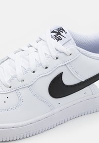 Biele kožené tenisky s čiernym logom Nike Swoosh, textúrovanou podrážkou, hrubými bielymi šnúrkami a polstrovaným jazykom s logom. Zaoblený dizajn špičky.