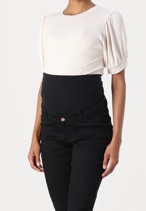 Zwangere persoon draagt een wit geribd T-shirt met korte mouwen en zwarte zwangerschapsbroek met een hoge, rekbare tailleband, staand tegen een effen achtergrond.