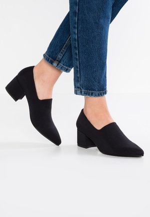 Sorte sko med lav blokhæl båret med blå, kortede jeans på en person, der står med den ene fod let hævet.