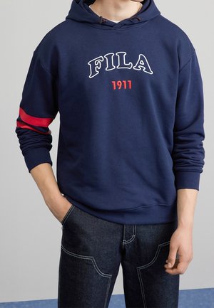 Osoba ubrana w granatową bluzę z kapturem FILA z czerwonym napisem "1911" i czerwonym paskiem na lewym rękawie, w połączeniu z ciemnymi jeansami z białym przeszyciem.
