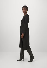 Abito nero a maniche lunghe con vita aderente e gonna svasata. Modella che indossa stivaletti neri con tacco a spillo al ginocchio; tessuto morbido e design sartoriale.