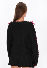 Schwarzer gestrickter Pullover mit lässiger Passform, der rosa Schleifen an den Schultern und eine strukturierte Oberfläche aufweist. Der Pullover hat lange Ärmel und einen runden Ausschnitt.