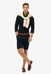 Dunkelblaues Hemd, beige Gürtel, dunkle Shorts, cremefarbene, grob gestrickte Pullover um den Hals gebunden, braune Slipper, selbstbewusste Pose.