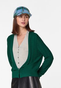 Grüner gestrickter Cardigan mit strukturiertem Design, offener Vorderseite und Knopfverschluss über einem grauen Oberteil. Das Model trägt eine blaue Karokappe.