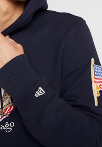 Marinblå hoodie med broderade grafiska motiv, inklusive en flaggdesign på ärmen och en logotyp på framsidan. Mjuk textur.