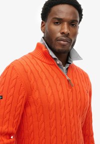 Pomarańczowy sweter w splot kablowy z wysokim kołnierzem i pół-zamkiem. Nosi się go na koszuli w paski z kołnierzykiem. Posiada prążkowane mankiety i dół.