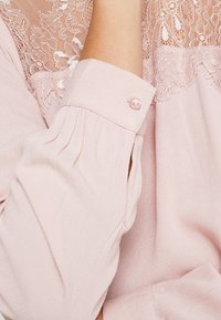 Blusa a maniche lunghe in rosa pallido, con una schiena in pizzo e polsini con bottone, dotata di un singolo bottone simile a una perla. Tessuto strutturato con pieghe sottili.