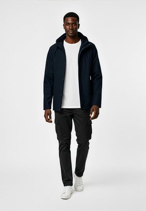 Marineblå vandtæt jakke med hætte, frontlås med trykknapper og sidelommer. Sammen med sorte cargo-bukser og hvide sneakers.
