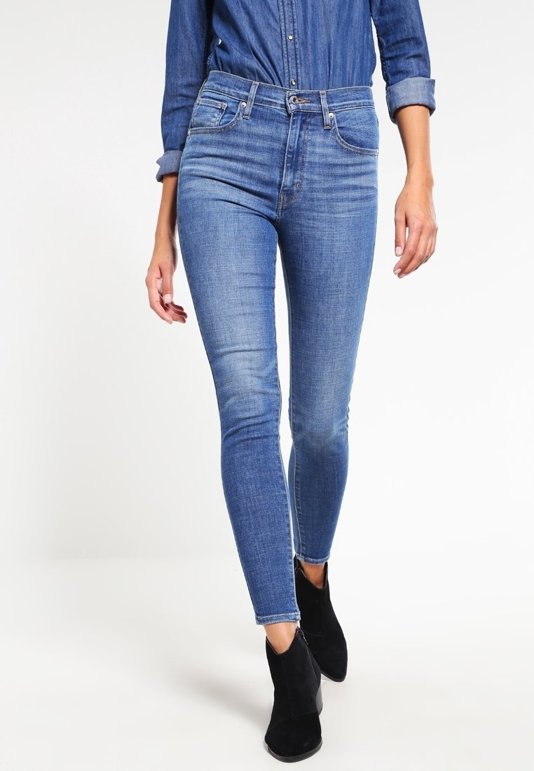 Femme portant un jean en denim bleu ajusté, une chemise en denim à manches longues retroussées et des bottines noires, marchant vers l'avant.