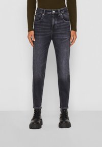 Jeans de mezclilla oscura con un corte slim, cinco bolsillos y cierre de botones. Presentan un sutil desgaste y costuras en contraste. Combinados con botas negras.