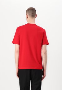 Personne aux cheveux courts et portant des boucles d'oreilles, vêtue d'un t-shirt rouge uni à manches courtes et d'un pantalon noir, debout dos à la caméra.