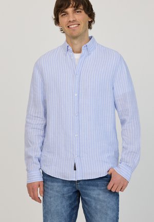 Mies hymyilee, hänellä on vaaleansininen ja valkoinen pystysuora raidallinen nappipaita valkoisen aluspaitansa päällä ja siniset farkut, seisoo yksiväristä taustaa vasten.