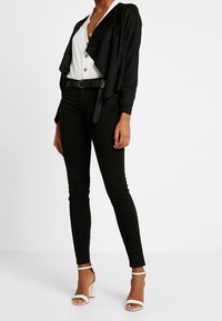 Sort cardigan med åbent frontstykke og lagdelt design, båret over en hvid skjorte med knapper. Kombineret med sorte skinny jeans og hvide sandaler med stropper.