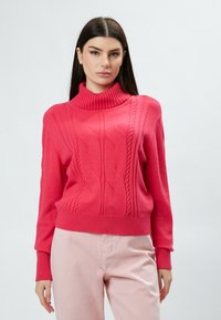 Pull en maille rose avec un col haut côtelé, présentant des motifs texturés et des manches allongées, associé à un pantalon rose clair.