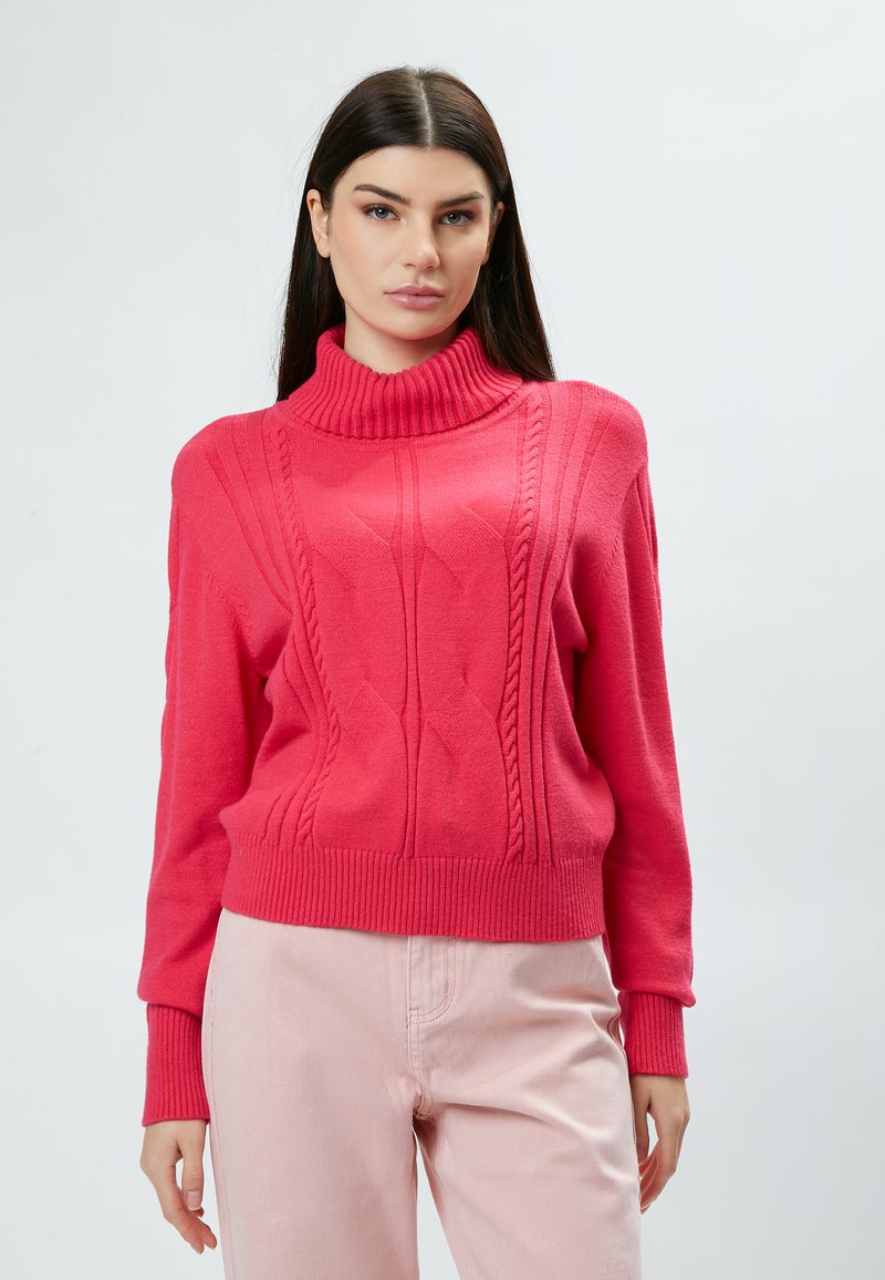 Pull en maille rose avec un col haut côtelé, présentant des motifs texturés et des manches allongées, associé à un pantalon rose clair.