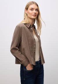 Camisa de corduroy marrón con botón frontal, cuello y mangas largas, combinada con una camiseta beige y jeans azul oscuro. Textura suave, corte relajado.