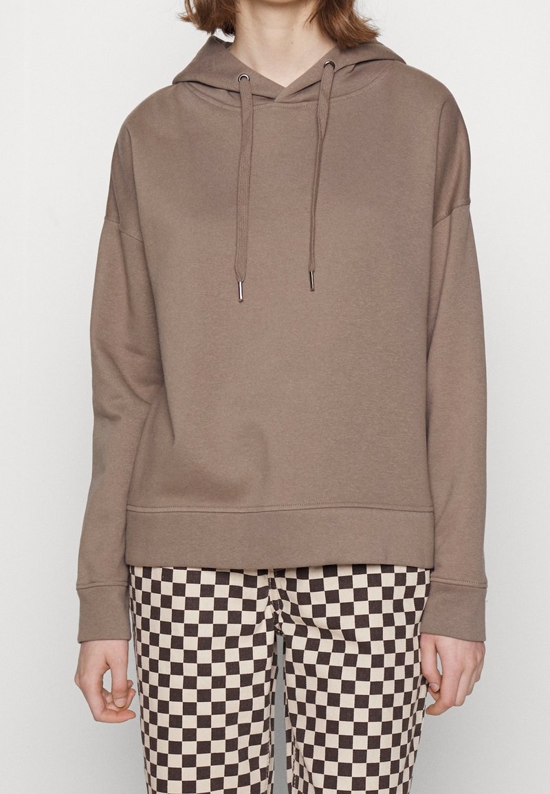 Brązowa bluza z kapturem o luźnym kroju, wyposażona w ściągacze z troczkami i ściągacze przy mankietach, zestawiona z czarno-kremowymi spodniami w kratę.