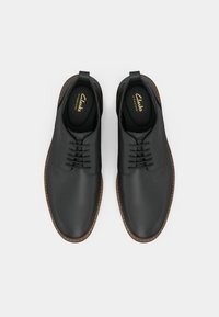 Pantofi din piele neagră cu un finisaj neted, având un design cu șireturi, tălpi din cauciuc maro și logo-ul "Clarks" în interiorul încălțămintei.