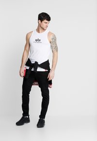 Hvit tanktop med logo, svarte jeans og svarte joggesko. En svart jakke knyttet rundt livet har røde tekstavsnitt.