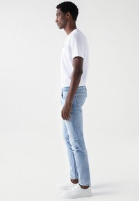 Jeans de color azul claro con un ajuste slim, llevados con una camiseta blanca y zapatillas blancas, mostrados de perfil contra un fondo liso.