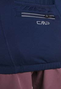 Gros plan sur un tissu bleu marine avec une petite poche zippée et un logo "CMP" blanc au-dessus d'un pantalon mauve.