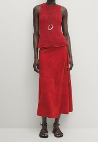 Haut rouge en tricot sans manches associé à une jupe portefeuille fluide en daim rouge. Comprend un collier rond métallique et des sandales marron.