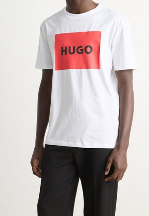 Man wearing white short-sleeve T-shirt with a red rectangle and black "HUGO" text, paired with black pants, against a plain background.