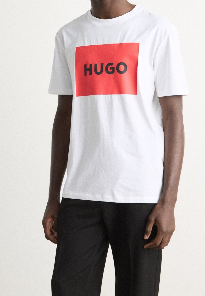 Hombre que lleva una camiseta blanca de manga corta con un rectángulo rojo y el texto negro "HUGO", combinado con pantalones negros, frente a un fondo liso.
